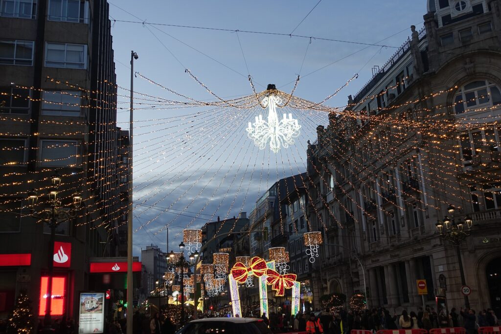 Dosel de luces navideñas con una lámpara de araña iluminada suspendida sobre una calle comercial de Vigo al anochecer