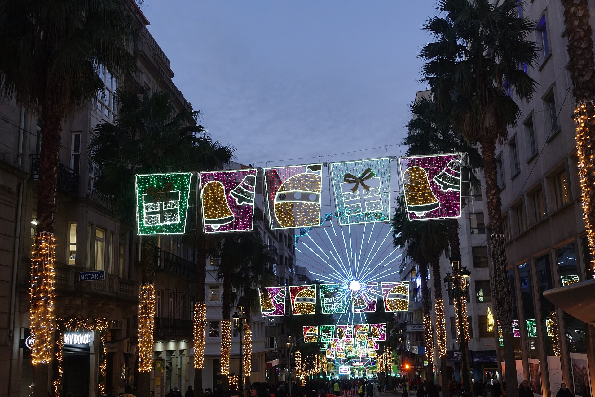 Paneles luminosos con campanas, regalos y bolas suspendidos sobre una calle de Vigo flanqueada por palmeras iluminadas, al anochecer