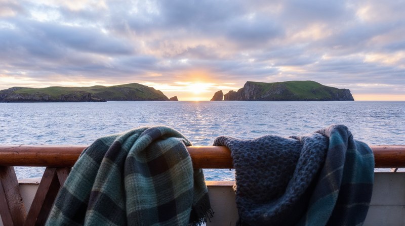 Imagen conceptual de unas islas verdes del Atlántico vistas desde un barco al atardecer de invierno, con bufandas de lana sobre la barandilla de madera