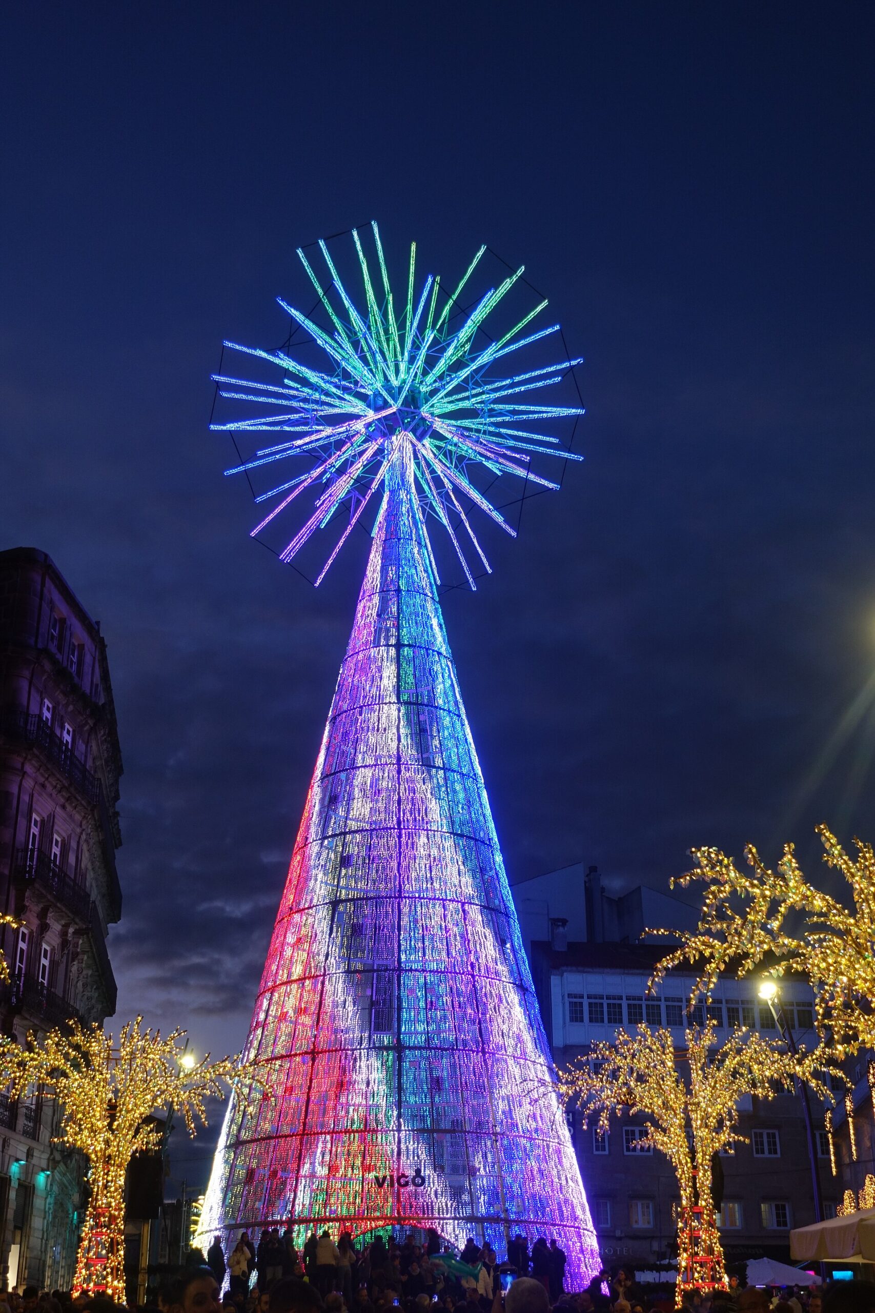 El árbol gigante de Navidad de la Porta do Sol de Vigo con patrones lumínicos multicolor, de noche, rodeado de árboles iluminados y con público a sus pies