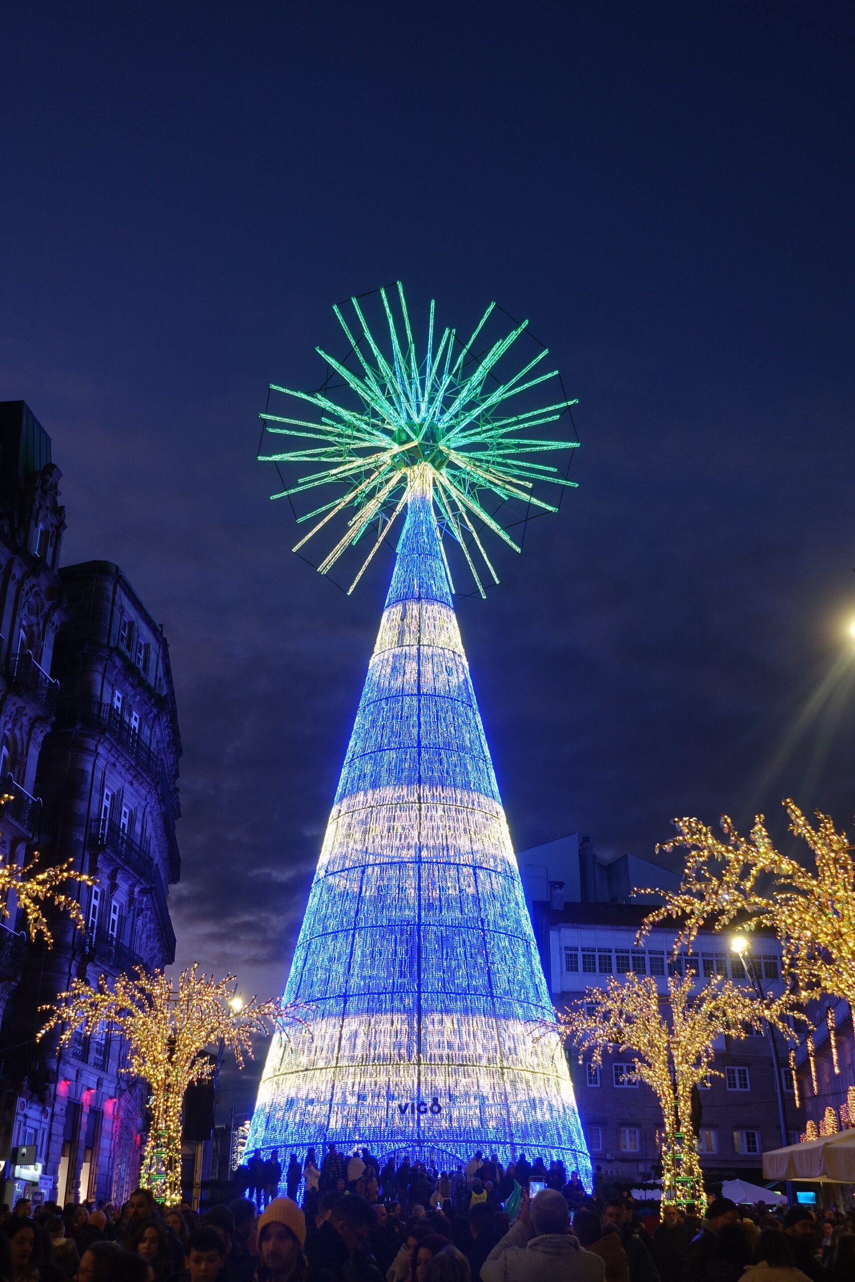 El árbol gigante de la Porta do Sol de Vigo iluminado en azul y blanco con su estrella verde, de noche, con público a sus pies