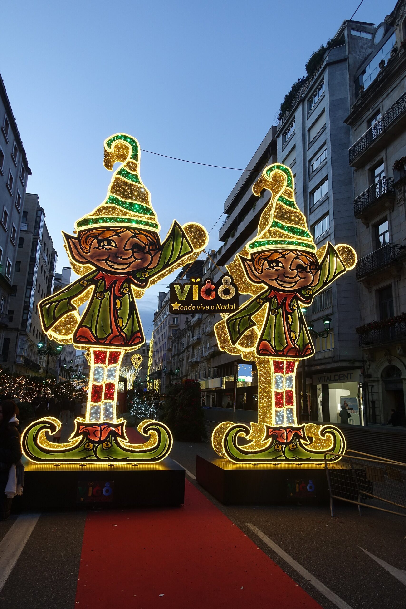 Dos figuras luminosas de elfos navideños en una calle del centro de Vigo al anochecer, con la señal de Vigo al fondo