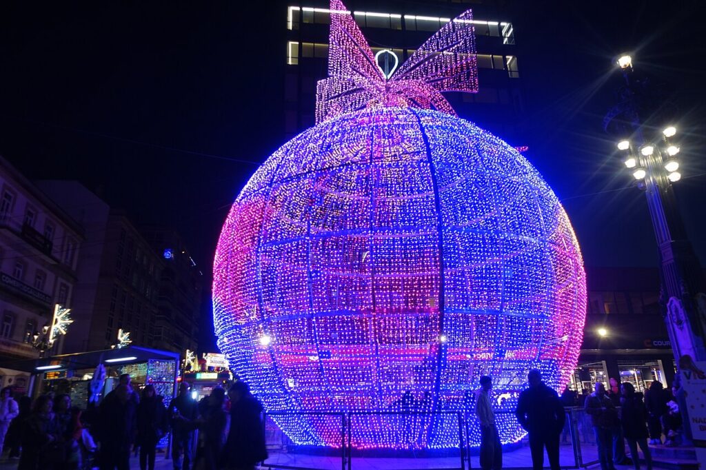Bola de Navidad gigante cubierta de luces rosas y azules con un lazo iluminado en la parte superior, de noche, con personas paseando a su alrededor