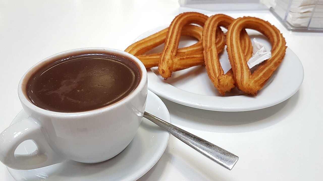 Taza de chocolate caliente espeso junto a un plato blanco con varios churros