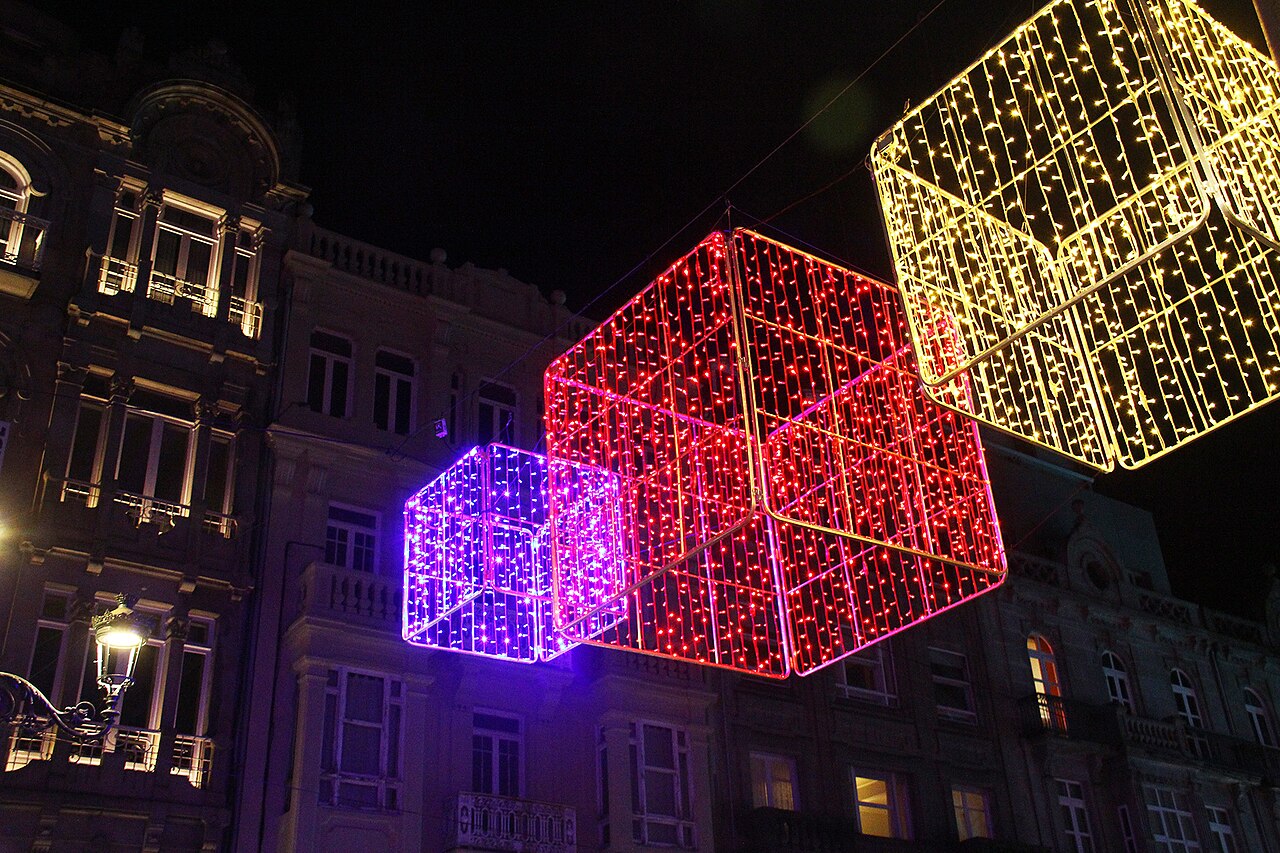 Tres cubos luminosos de colores rojo, morado y amarillo suspendidos sobre una calle de noche, con edificios iluminados al fondo