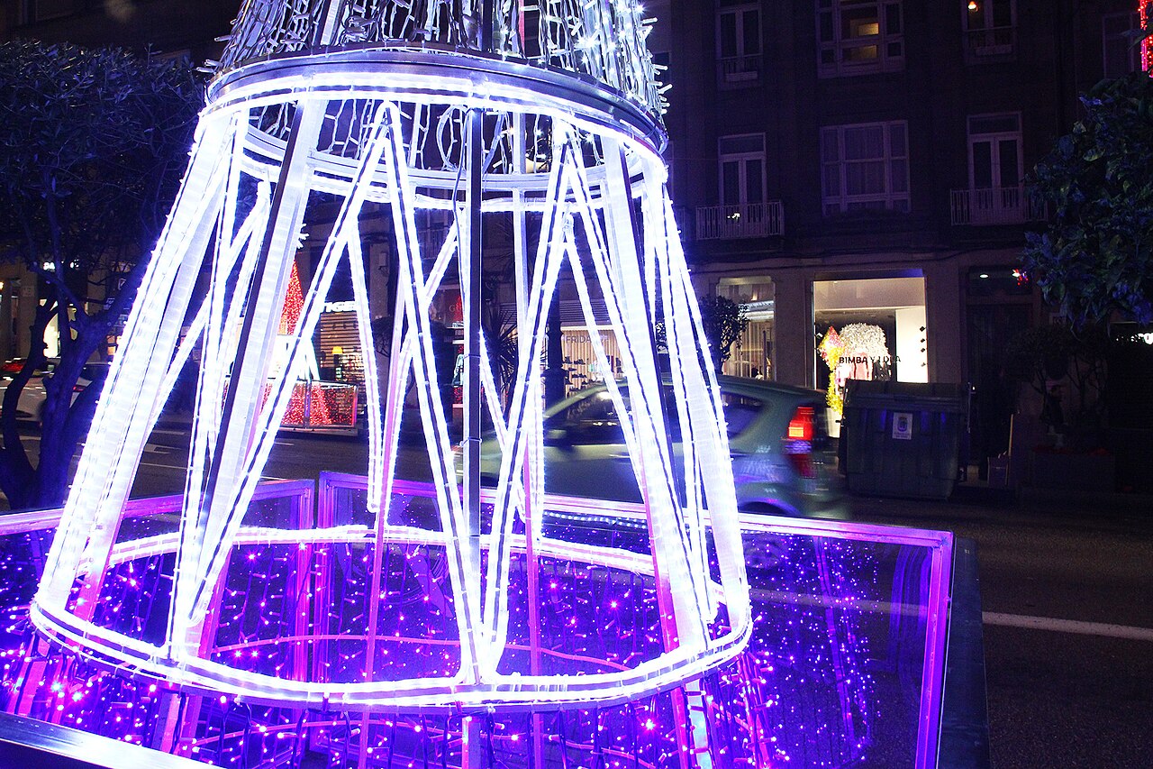 Estructura luminosa blanca con forma de farol de varios metros en una calle de noche, con tiendas iluminadas al fondo