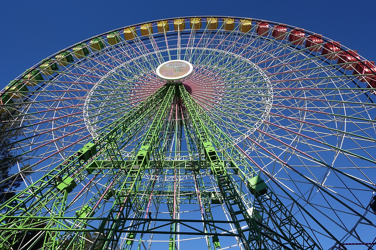 Noria de gran tamaño con estructura verde y cabinas amarillas y rojas, vista desde abajo con cielo azul despejado